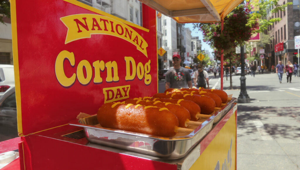 「NATIONAL Corn Dog DAY」と書かれた屋台に、マスタードがかかったコーンドッグが並び、街を歩く人々が背景にいる。